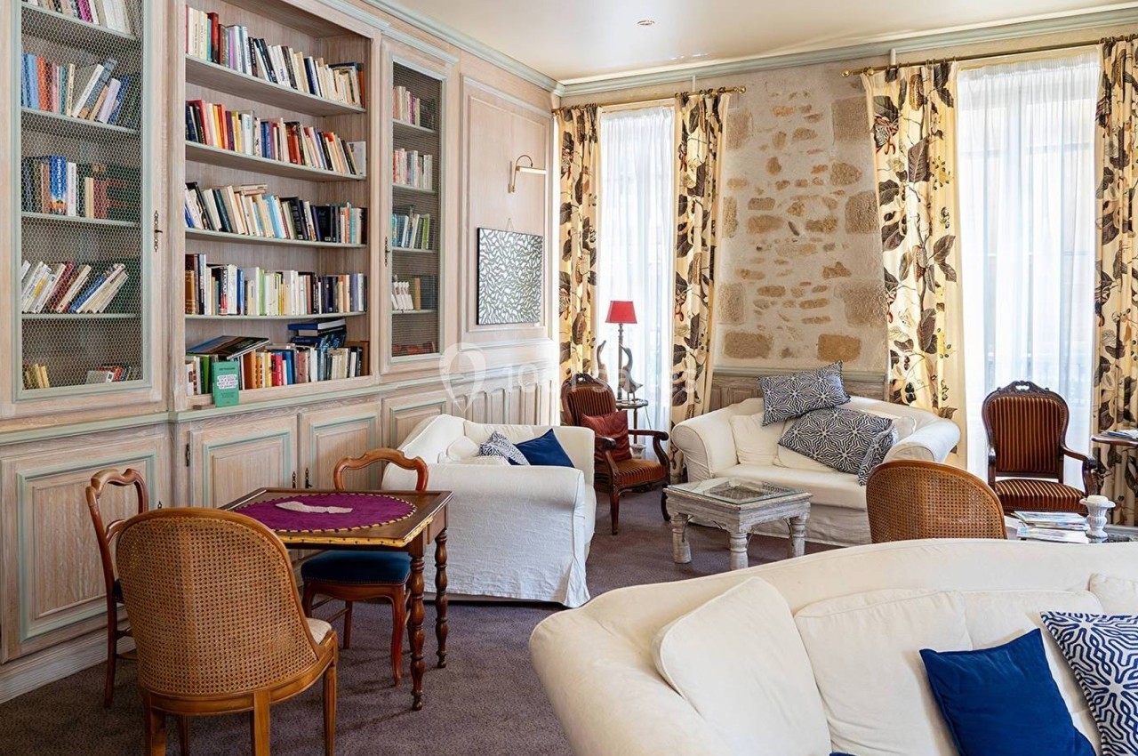 Salon lumineux avec bibliothèque en bois, fauteuils et canapés, murs en pierre et grandes fenêtres avec rideaux fleuris.