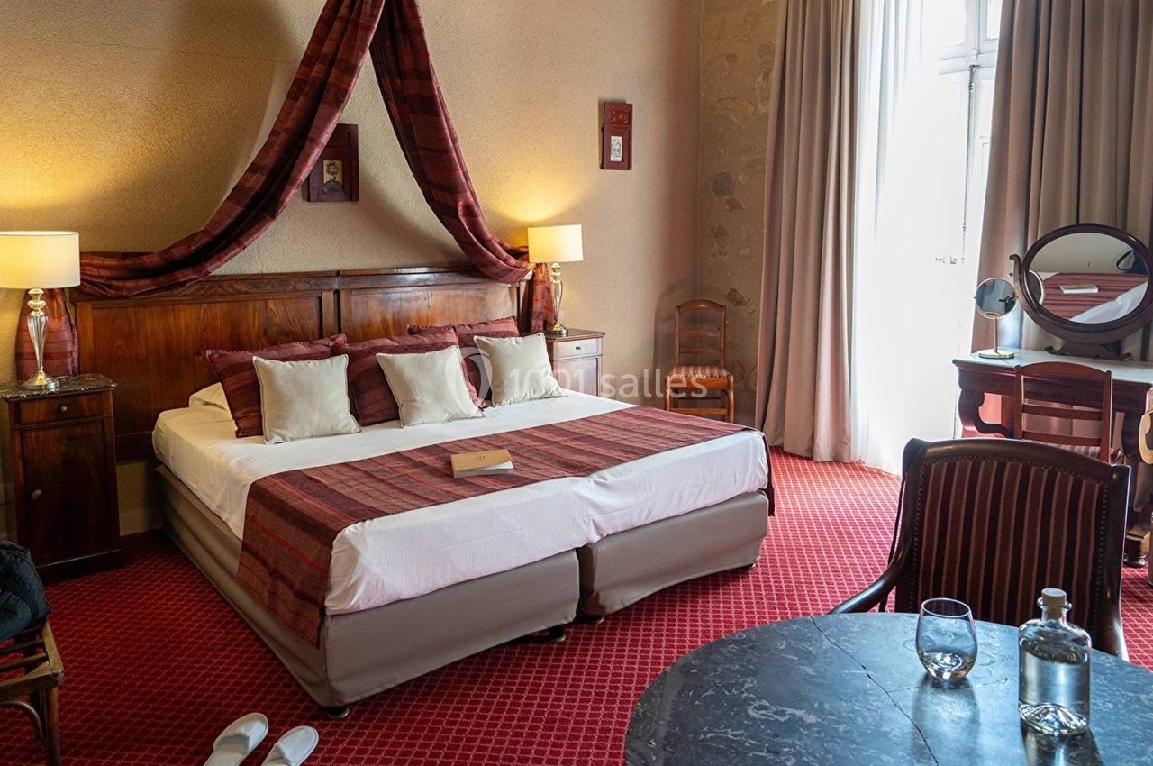 Chambre d'hôtel avec lit double, décorée de tons rouges, équipée de lampes, d'une coiffeuse et d'une table avec carafe d'eau.