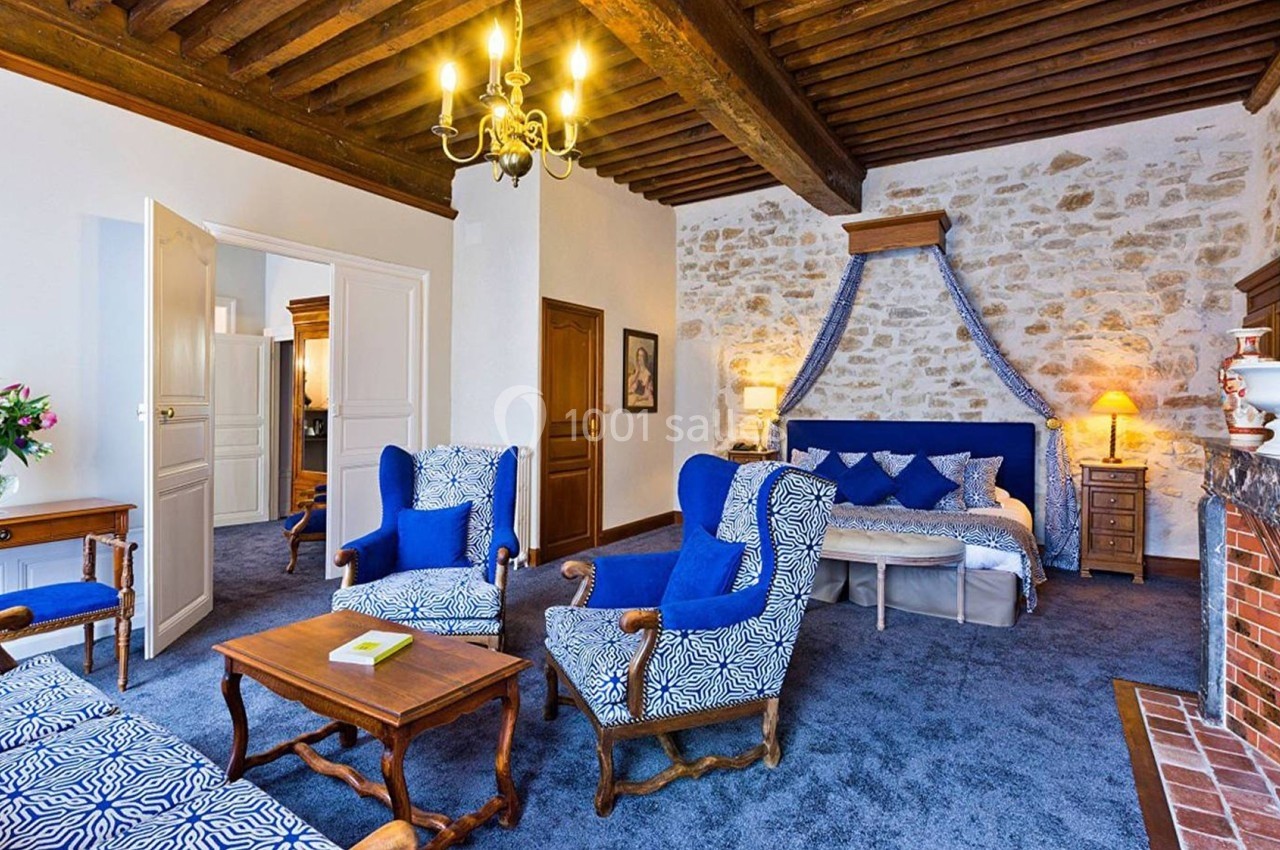 Chambre spacieuse avec murs en pierre, plafond à poutres apparentes, fauteuils bleus et lit à baldaquin.