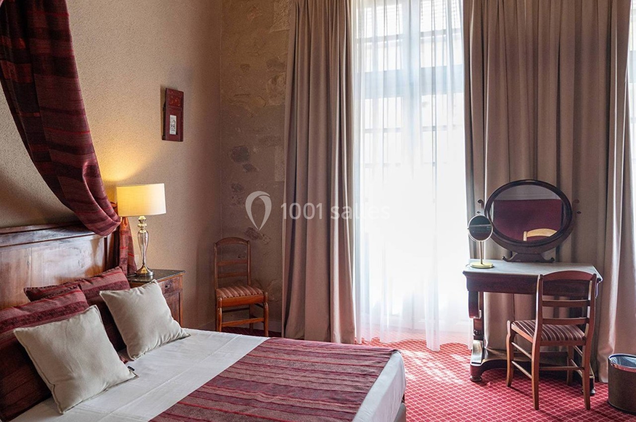Chambre lumineuse avec lit double, bureau avec miroir, chaises en bois et rideaux laissant passer la lumière naturelle.