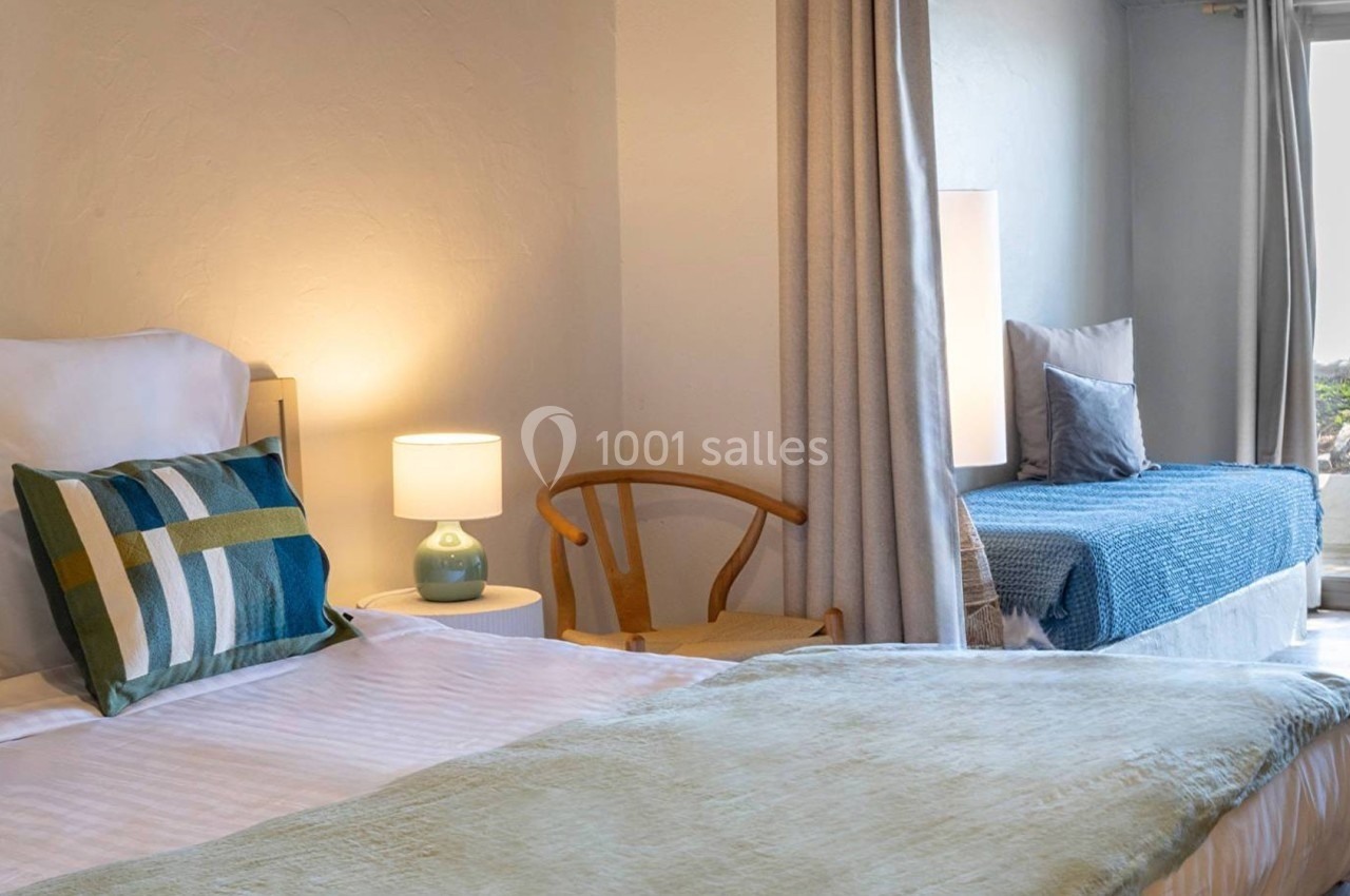 Chambre lumineuse avec lit double, lit d'appoint, fauteuil en bois, lampe de chevet et accès à une terrasse.