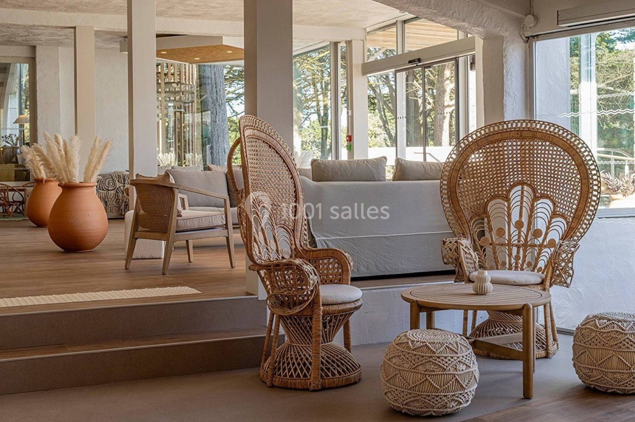 Espace intérieur lumineux avec fauteuils en rotin, table basse, poufs tressés et grande jarre décorative en arrière-plan.