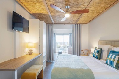 Piscine extérieure entourée de chaises longues et de tables, avec des bâtiments blancs et des arbres en arrière-plan.