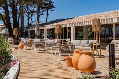 Piscine extérieure entourée de chaises longues et de tables, avec des bâtiments blancs et des arbres en arrière-plan.