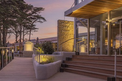 Piscine extérieure entourée de chaises longues et de tables, avec des bâtiments blancs et des arbres en arrière-plan.