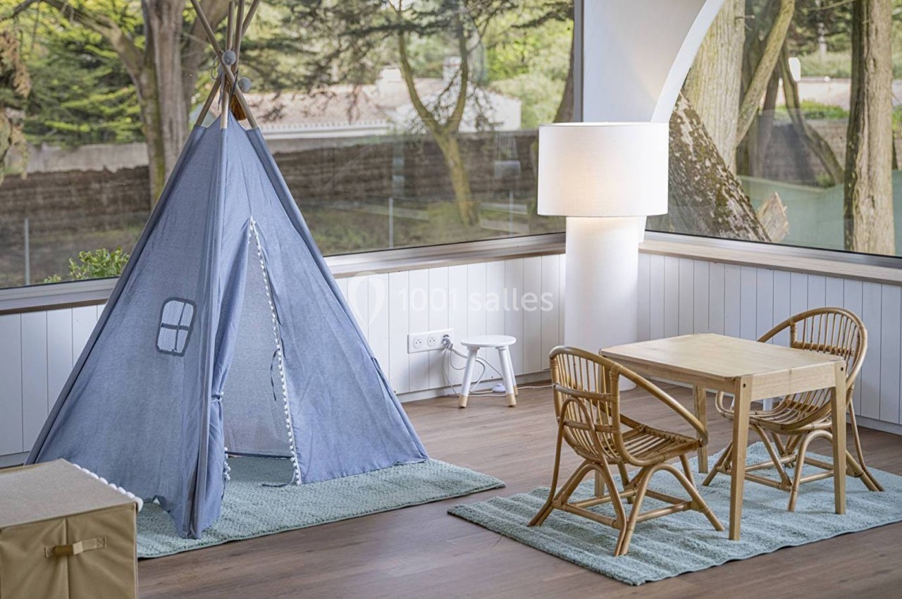 Tente bleue pour enfants et mobilier en rotin dans une pièce lumineuse avec vue sur un jardin boisé.