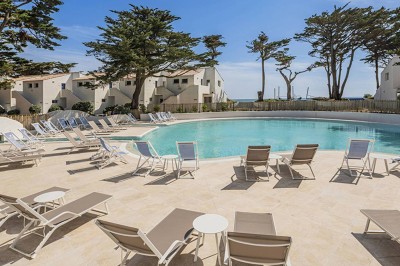 Piscine extérieure entourée de chaises longues et de tables, avec des bâtiments blancs et des arbres en arrière-plan.