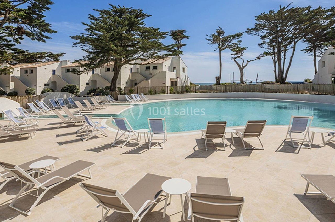Piscine extérieure entourée de chaises longues et de tables, avec des bâtiments blancs et des arbres en arrière-plan.