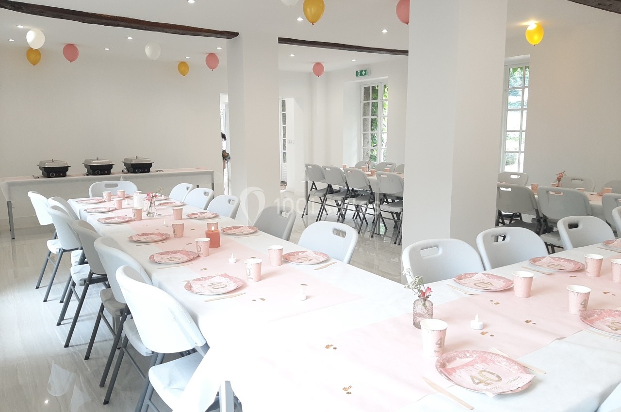Salle lumineuse décorée pour une réception, avec tables dressées de nappes blanches et roses, et ballons suspendus.