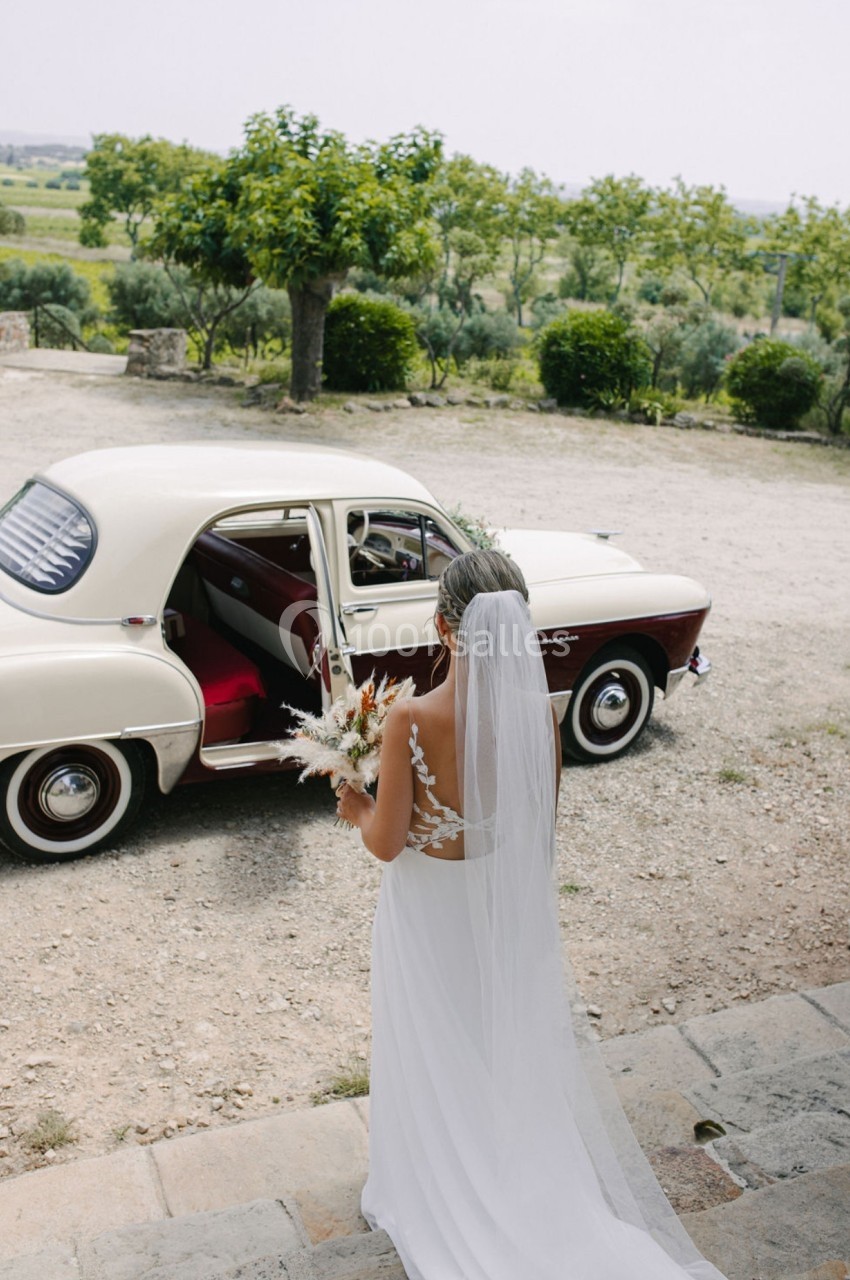 Une mariée en robe blanche tient un bouquet et regarde une voiture ancienne beige garée dans un cadre champêtre.