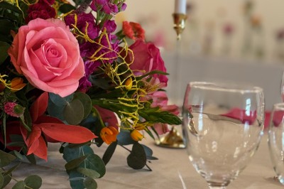 Centre de table floral coloré avec des roses et des verres disposés sur une nappe blanche, bougie en arrière-plan.