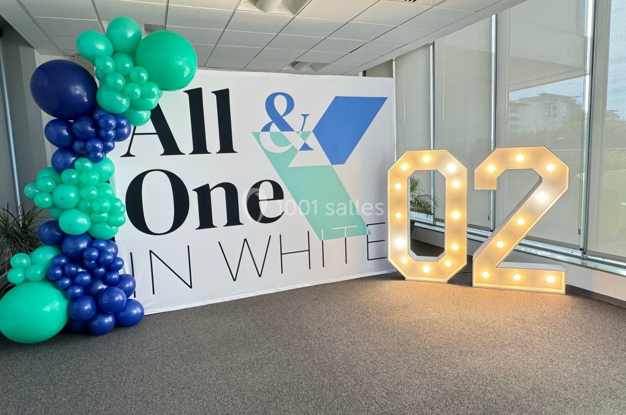 Mur décoratif avec ballons verts et bleus, panneau ’All & One in White’ et chiffre lumineux ’02’ dans un espace intérieur.