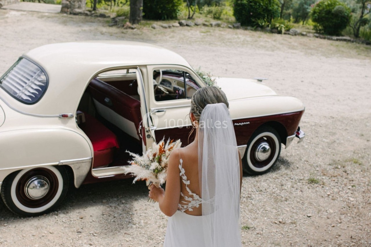 Une mariée en robe blanche et voile tient un bouquet devant une voiture ancienne beige et bordeaux, portière ouverte.
