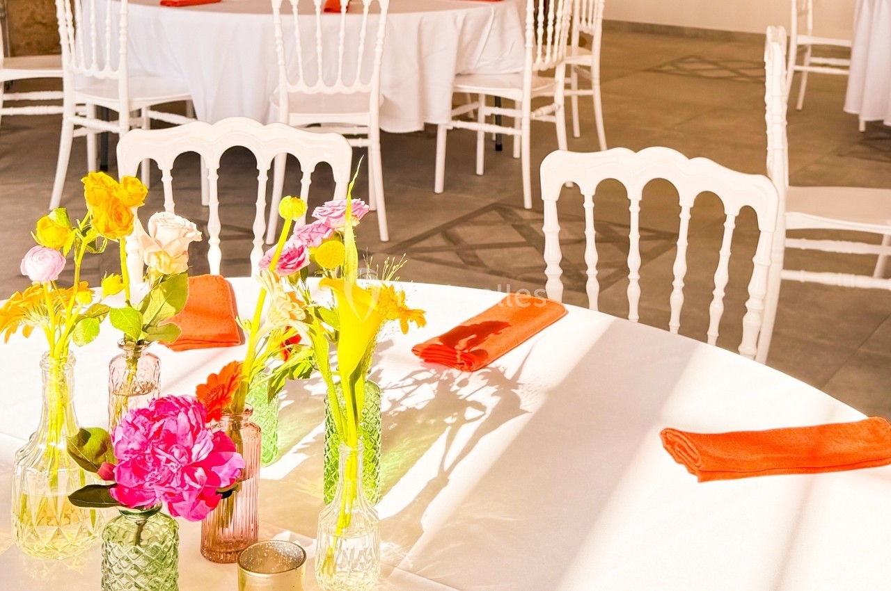 Table ronde décorée de fleurs colorées et de serviettes orange, entourée de chaises blanches dans une salle lumineuse.