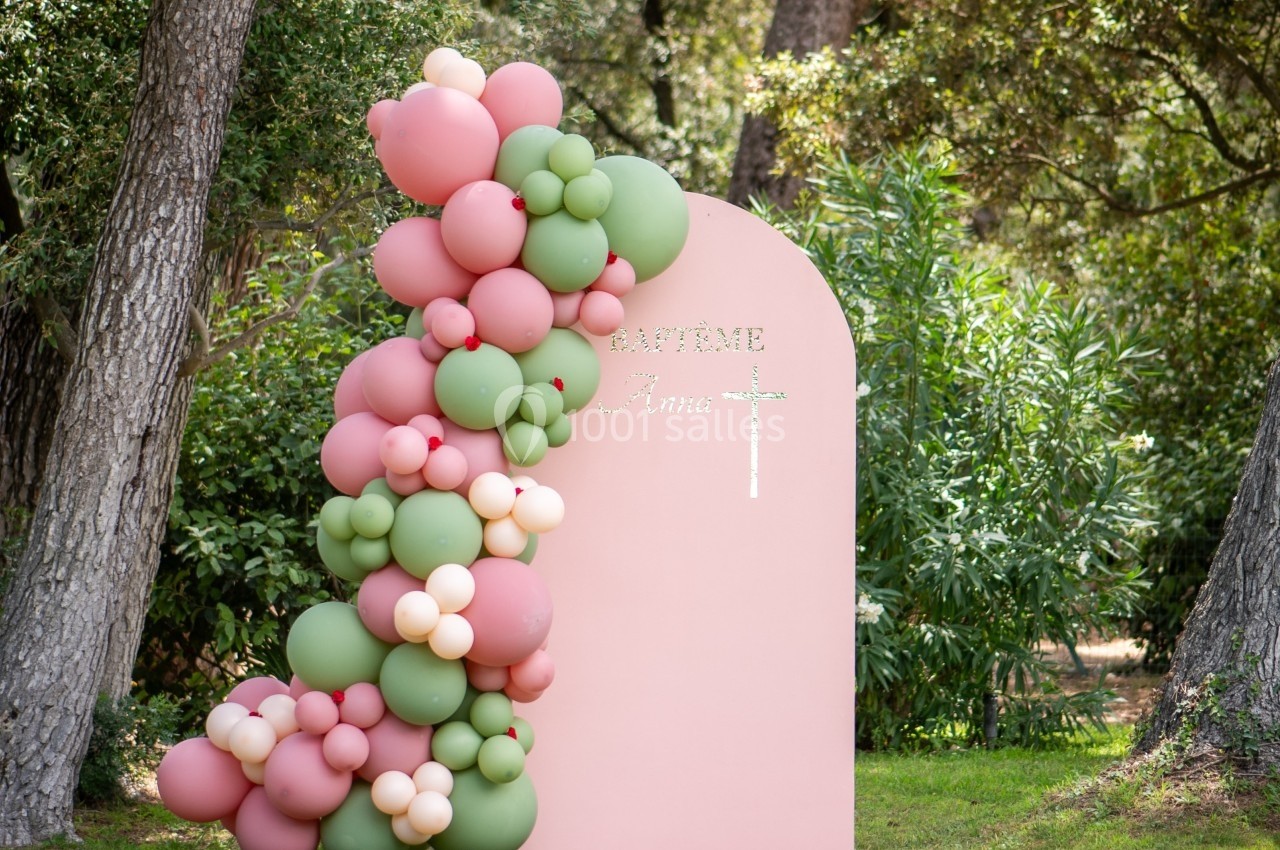 Décoration de baptême avec arche de ballons roses et verts devant un panneau rose dans un cadre boisé.