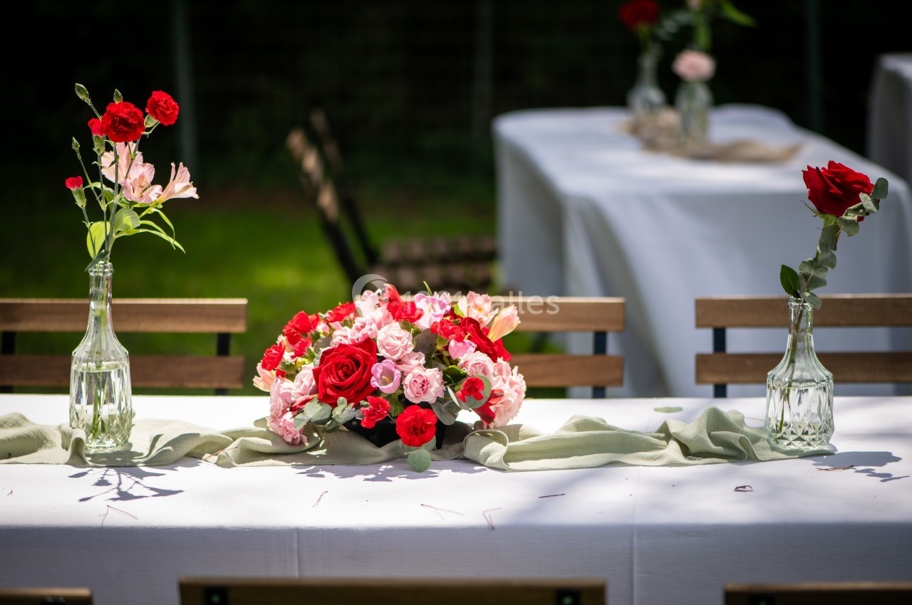 Bouquet de fleurs rouges et roses sur une table blanche décorée, entouré de vases et de chaises en bois.