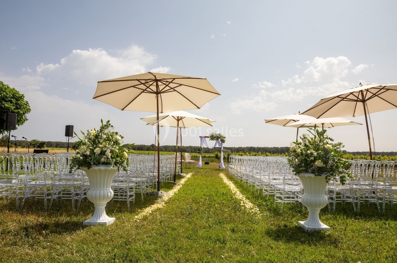 Allée centrale décorée de fleurs blanches menant à une arche de mariage, entourée de chaises et de parasols en plein air.
