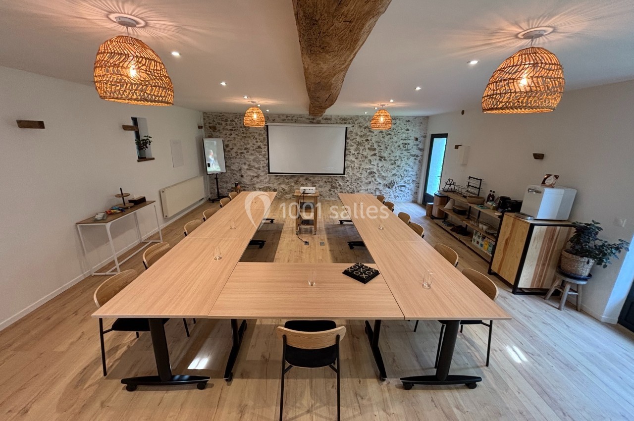 Salle de réunion lumineuse avec grande table en U, chaises, projecteur, murs en pierre et éclairage suspendu.