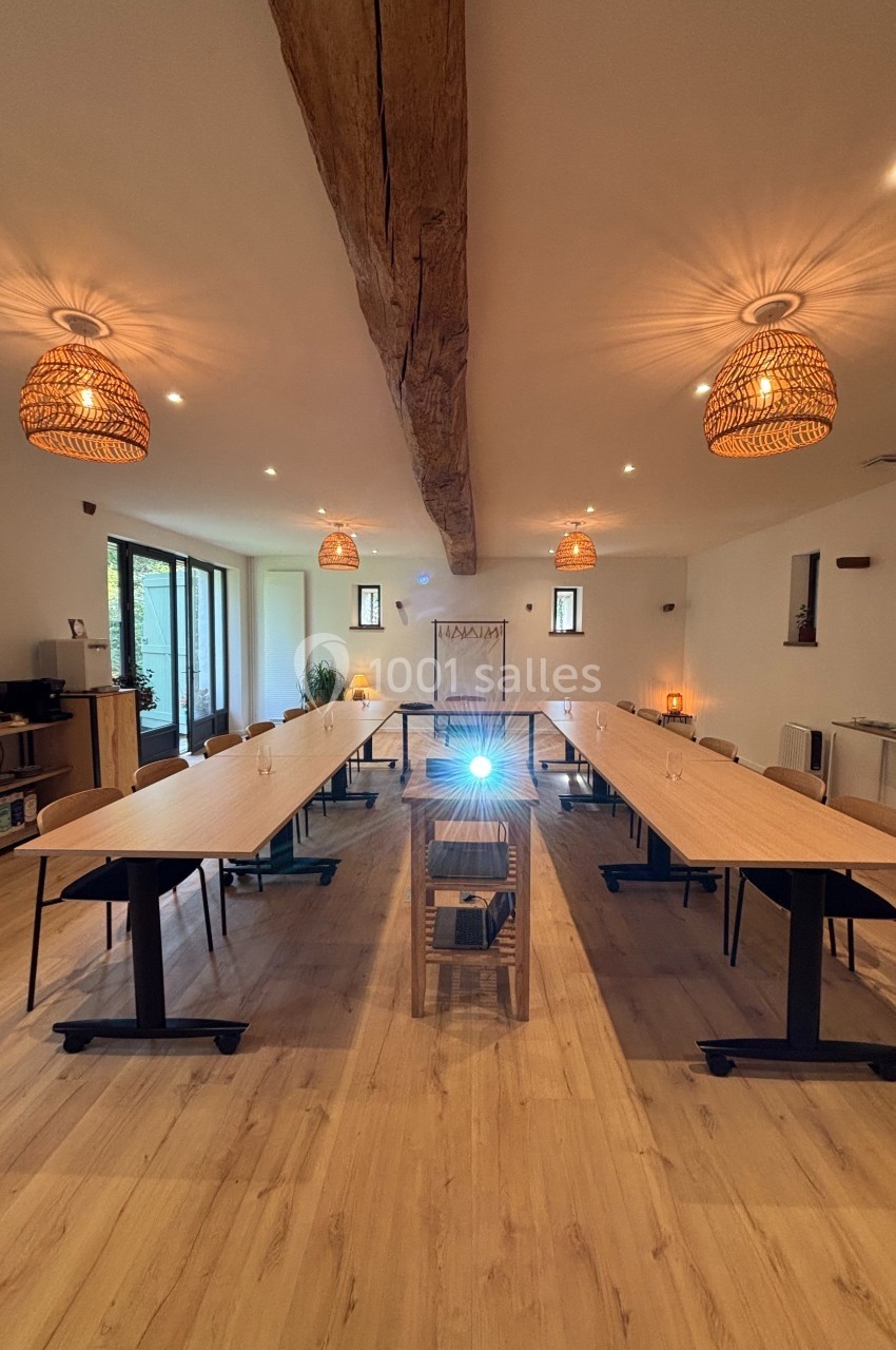 Salle de réunion lumineuse avec tables disposées en U, projecteur allumé et poutre apparente au plafond.