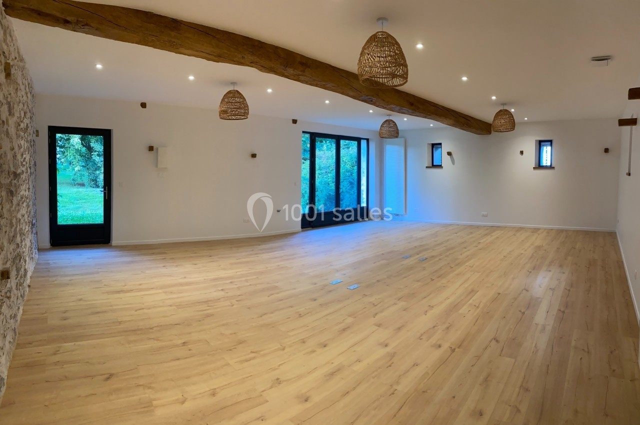 Salle spacieuse avec parquet clair, murs blancs, poutres apparentes et grandes baies vitrées donnant sur un jardin.