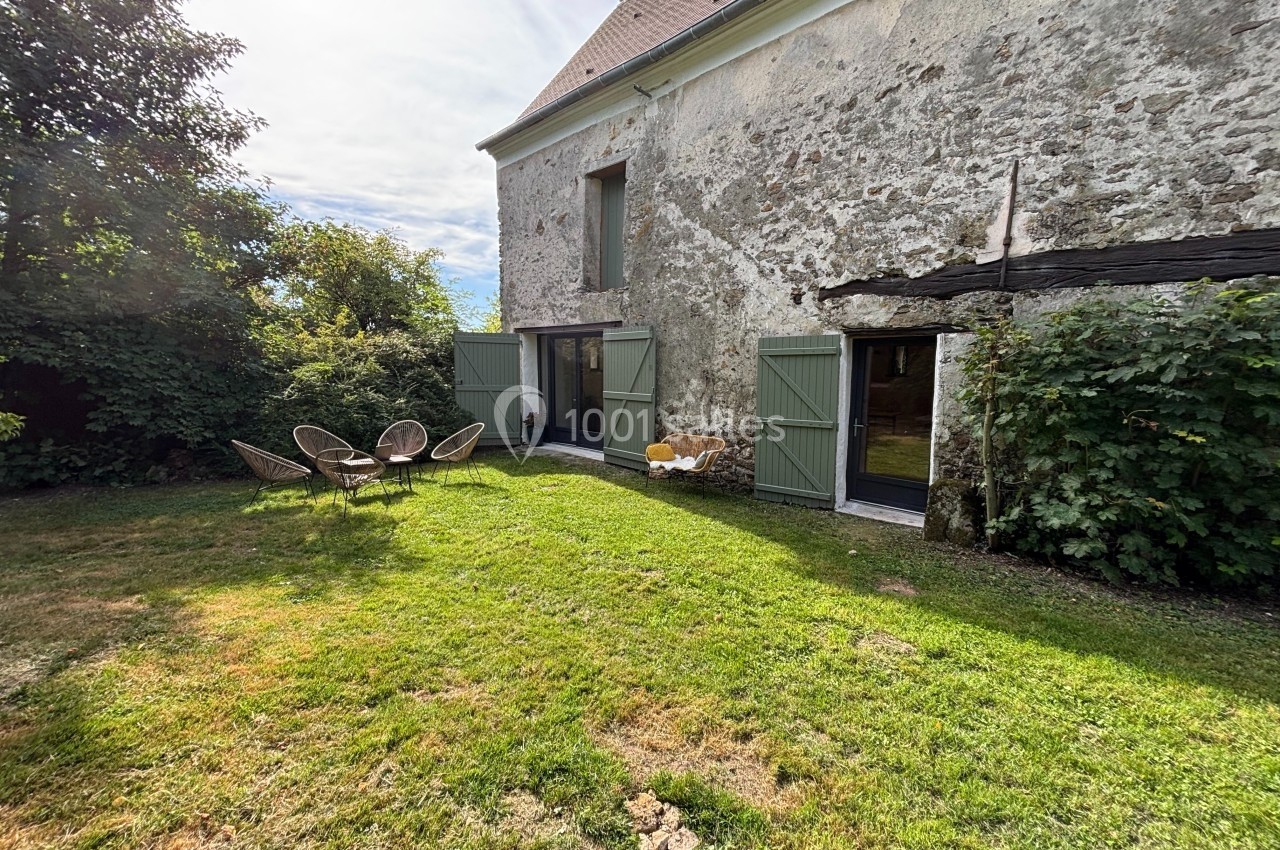 Façade en pierre d'une maison avec portes vertes, jardin herbeux, mobilier d'extérieur et végétation environnante.