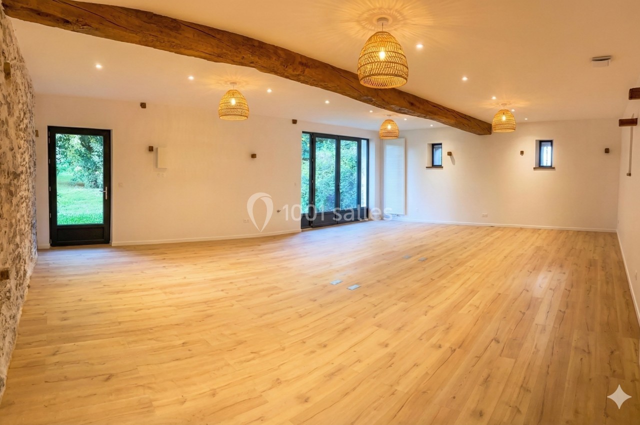Salle lumineuse avec parquet en bois clair, murs blancs, poutres apparentes et grandes fenêtres donnant sur un jardin.