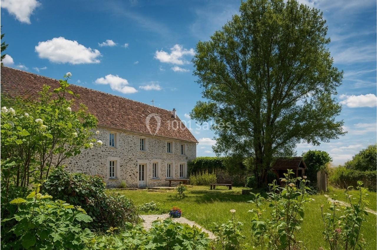 Maison en pierre avec toit en tuiles, entourée d'un jardin verdoyant et d'arbres sous un ciel bleu.