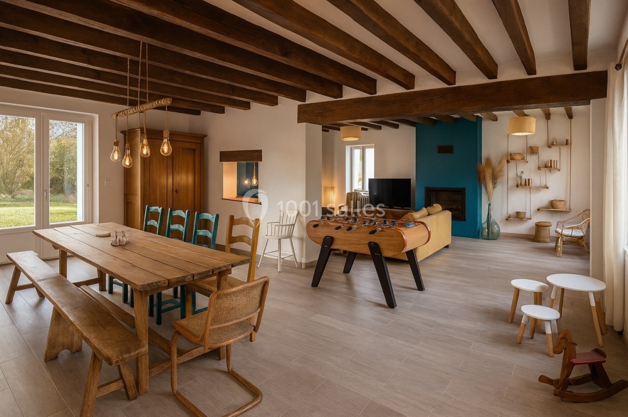 Salle de séjour lumineuse avec table en bois, baby-foot, canapé jaune, poutres apparentes et vue sur un jardin.