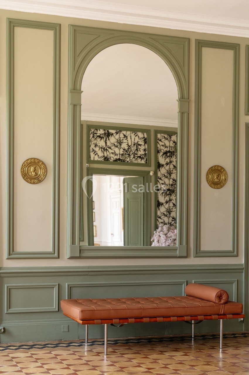 Banc en cuir marron devant un grand miroir encadré, dans une pièce aux murs verts ornés de moulures et médaillons dorés.