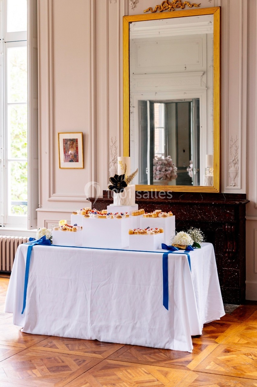 Table décorée avec une pièce montée blanche, entourée de fleurs et rubans bleus, dans une salle élégante avec miroir doré.