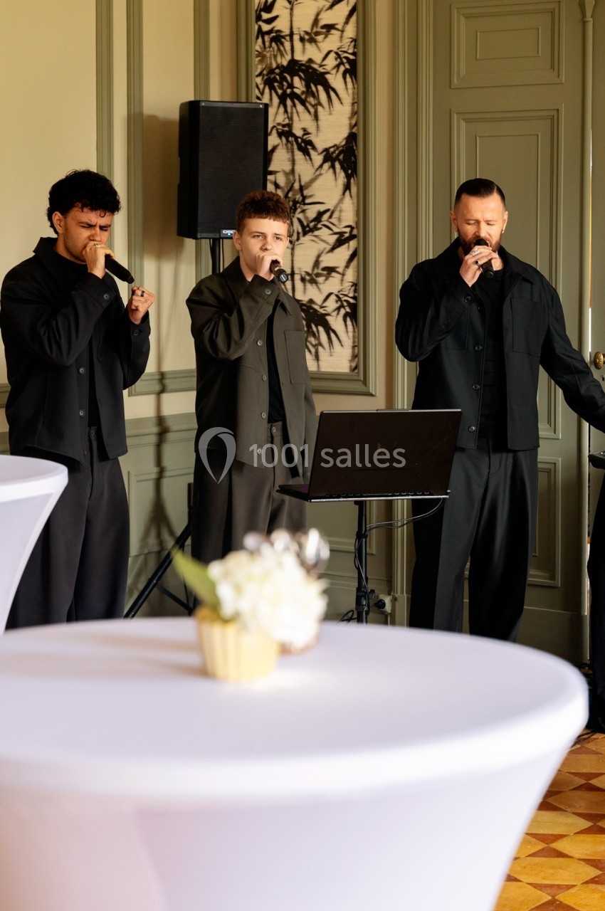 Trois chanteurs habillés en noir interprètent une chanson dans une salle élégante avec des tables en premier plan.