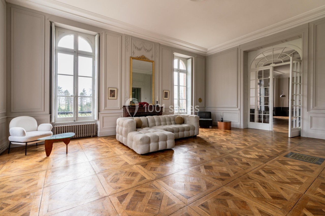 Salon lumineux avec parquet en bois, canapé beige, fauteuil, table basse et grandes fenêtres donnant sur l'extérieur.
