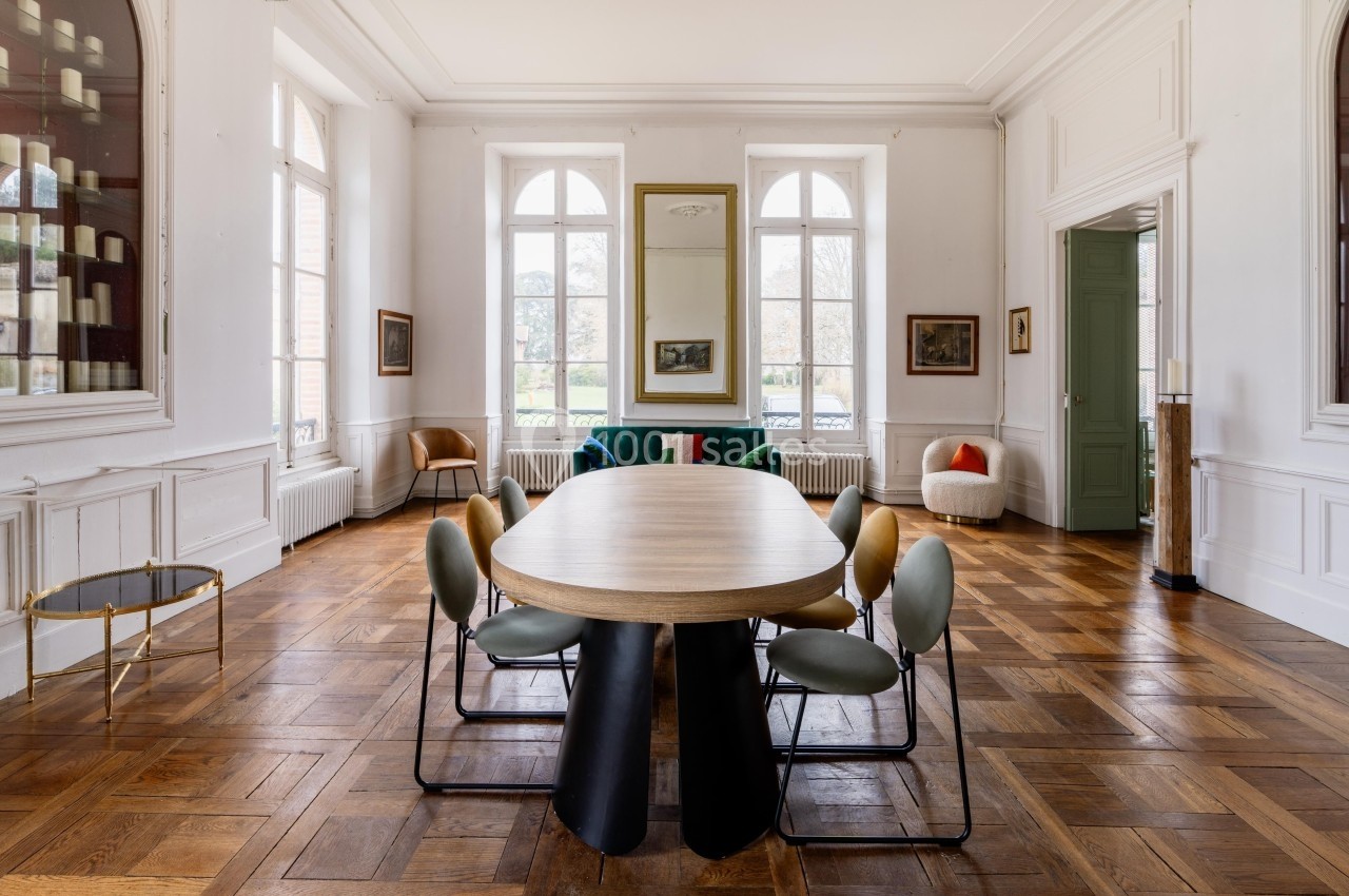 Salle à manger lumineuse avec table ovale en bois, chaises modernes et parquet en chevrons, entourée de grandes fenêtres.