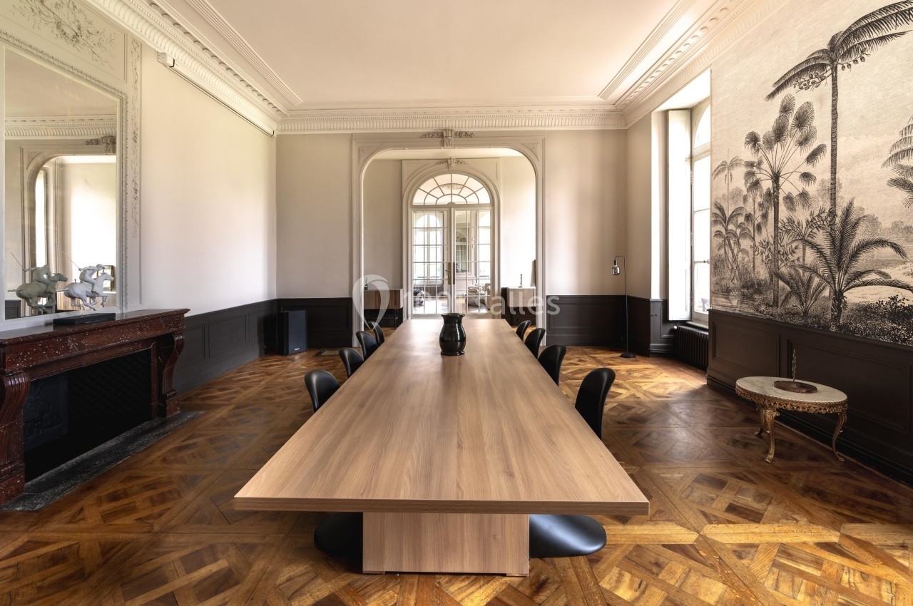 Salle de réunion lumineuse avec une grande table en bois, des chaises noires, parquet en chevrons et fresque murale…