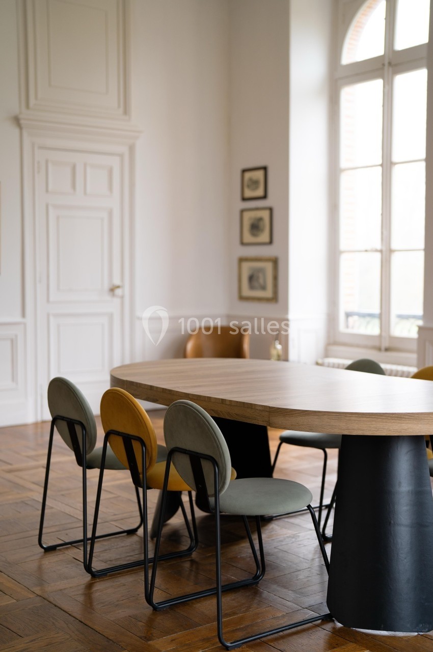 Salle lumineuse avec une grande table en bois, chaises modernes et parquet, près de fenêtres hautes et murs blancs.