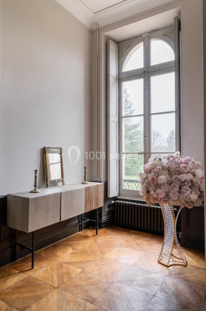 Console moderne avec bougeoirs et miroir, près d'une fenêtre cintrée, bouquet de fleurs sur un pied en verre à droite.