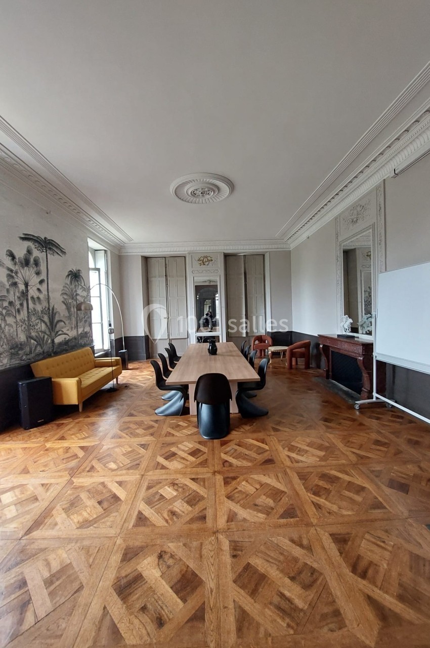 Salle de réunion lumineuse avec parquet en bois, grande table, chaises noires, canapé jaune et décor mural tropical.