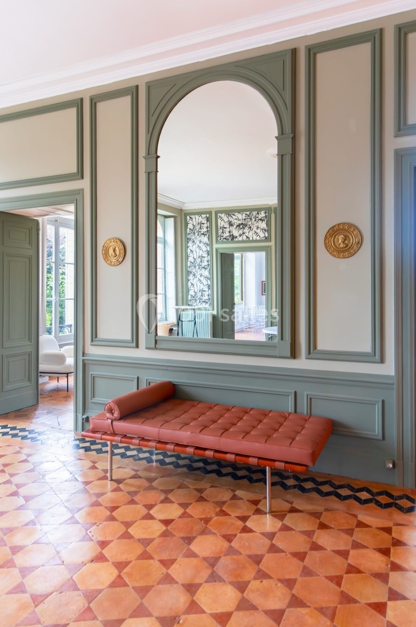 Banc en cuir marron devant un grand miroir encadré de moulures vertes, dans une pièce lumineuse au sol en carreaux.
