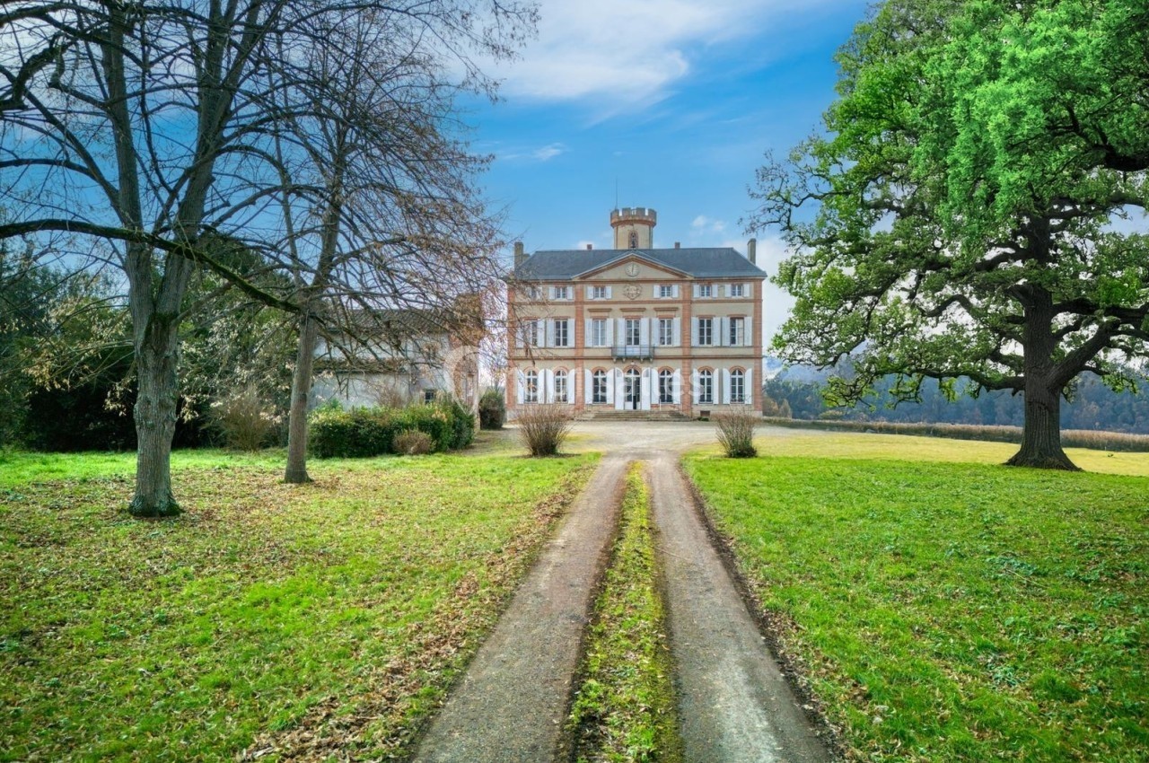Allée bordée de pelouse menant à un grand manoir en pierre entouré d'arbres, sous un ciel dégagé.