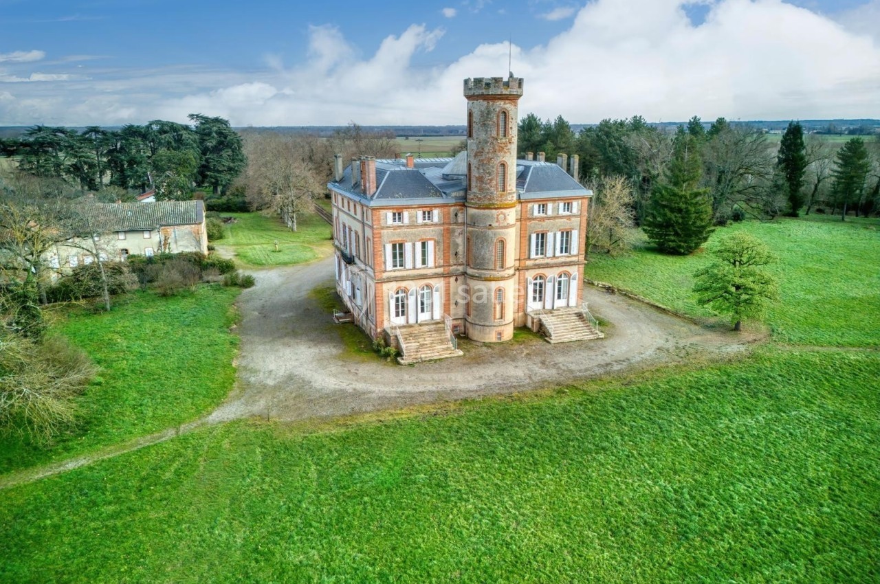 Château en briques rouges avec une tour centrale, entouré de pelouses et d'arbres, sous un ciel partiellement nuageux.