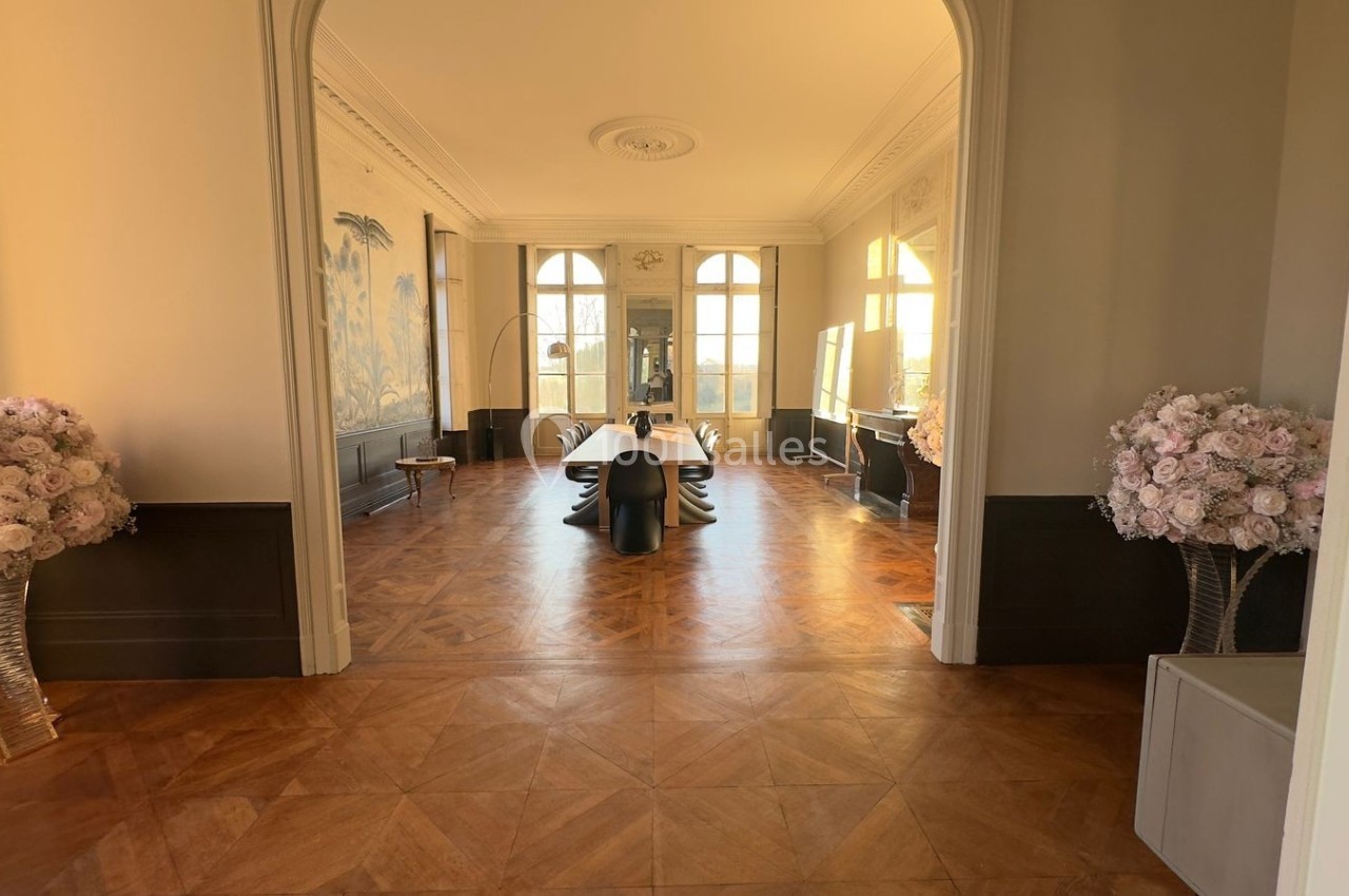 Salle lumineuse avec parquet en bois, table noire centrale, murs ornés et bouquets de fleurs roses sur les côtés.