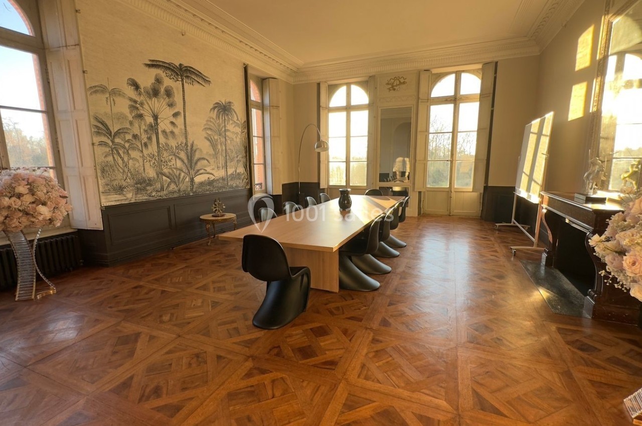 Salle de réunion lumineuse avec une grande table en bois, chaises noires, parquet en chevrons et décor mural végétal.