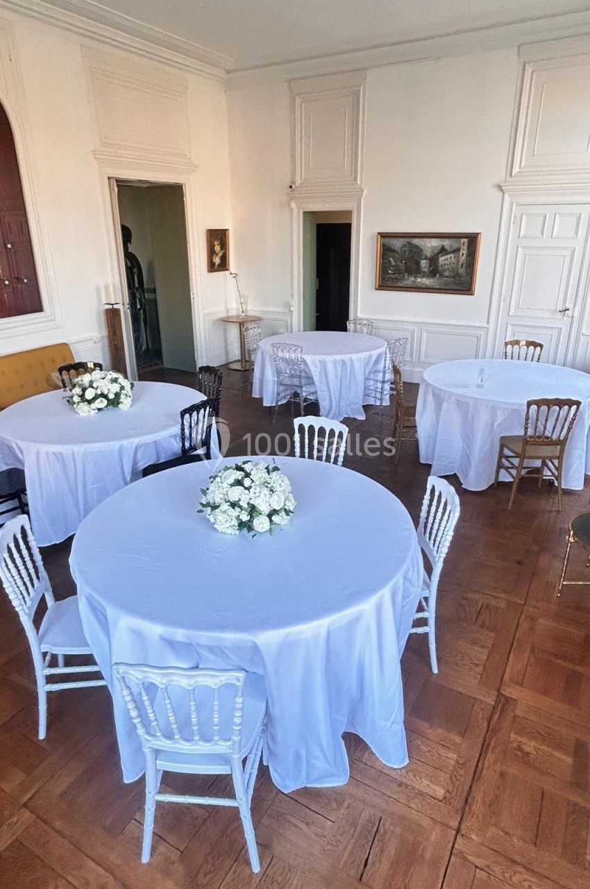 Salle avec parquet, tables rondes nappées de blanc, chaises variées et décorations florales au centre des tables.