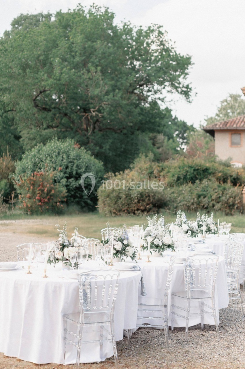 Tables rondes décorées de nappes blanches et de fleurs, disposées en extérieur près d'arbres et d'un bâtiment.