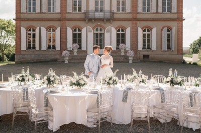 Un couple en tenue de mariage se tient devant une grande table décorée, avec un manoir en arrière-plan.