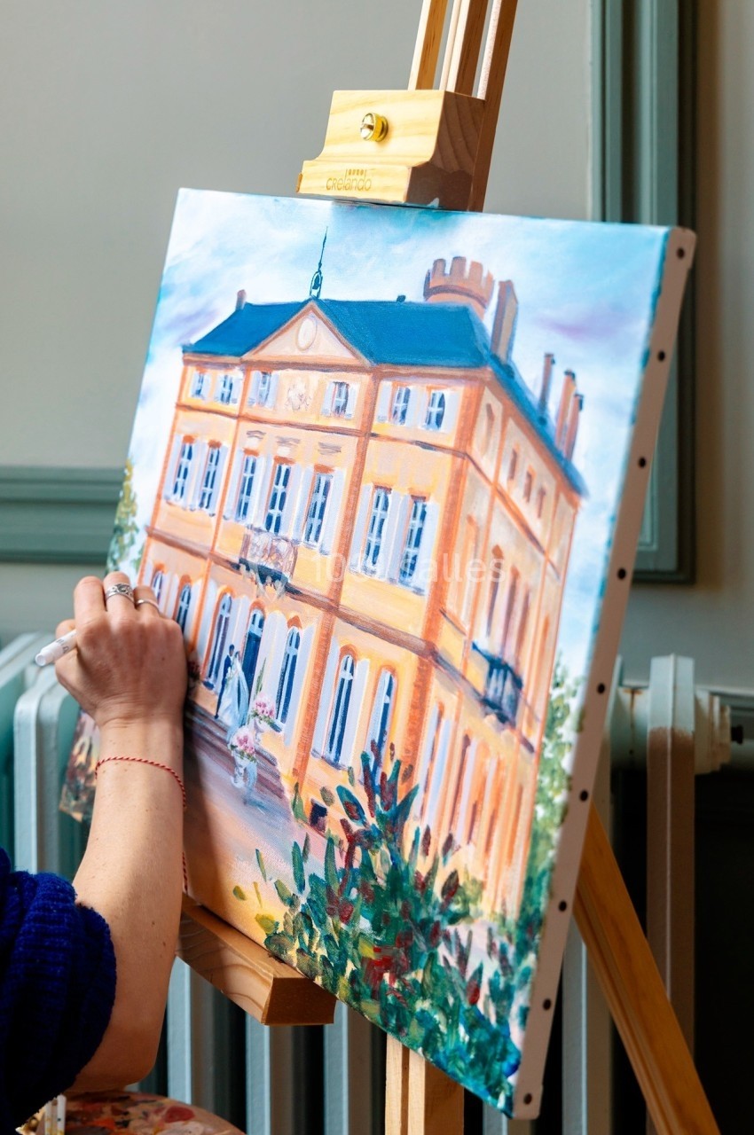 Une personne peint un tableau représentant un bâtiment jaune avec des détails architecturaux, posé sur un chevalet.