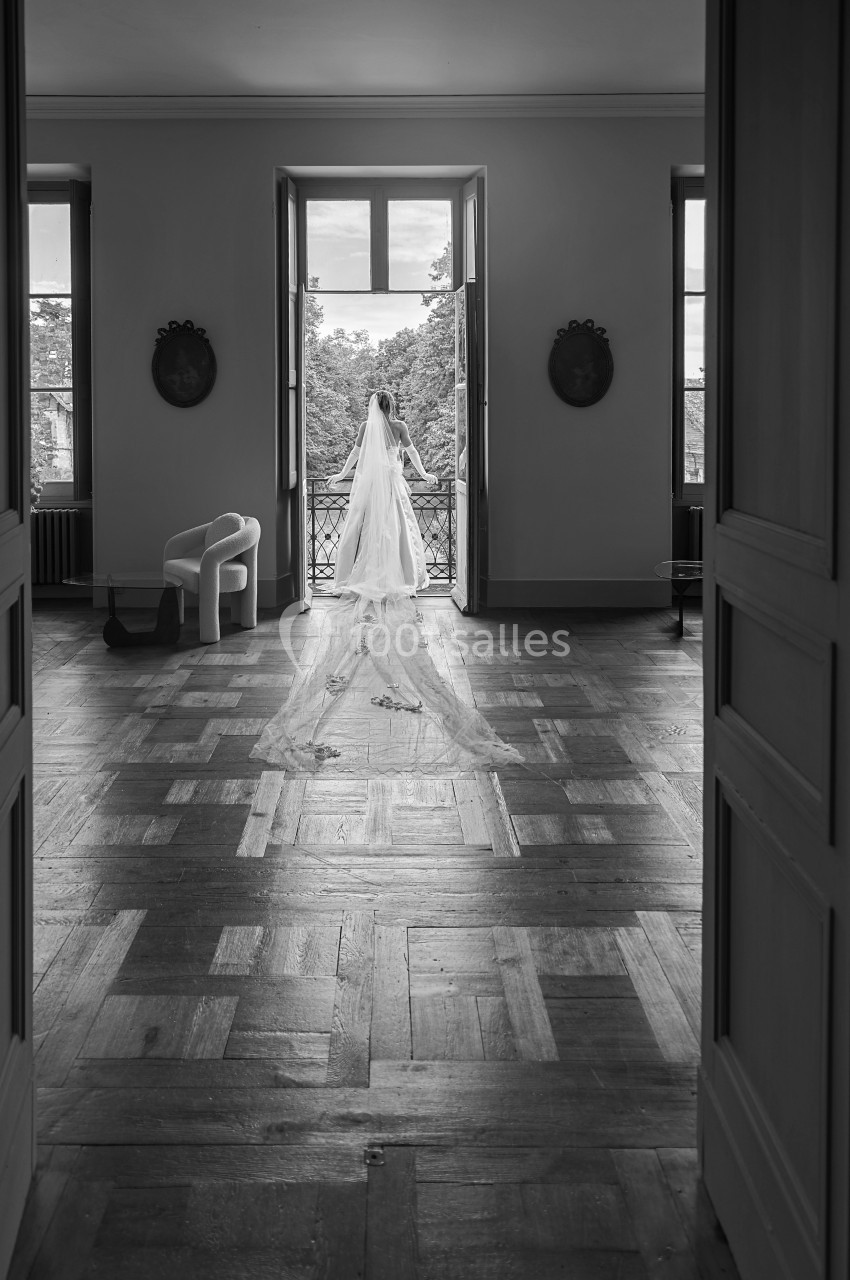 Une robe de mariée avec un long voile est posée dans une pièce lumineuse avec parquet, face à des portes-fenêtres ouvertes.