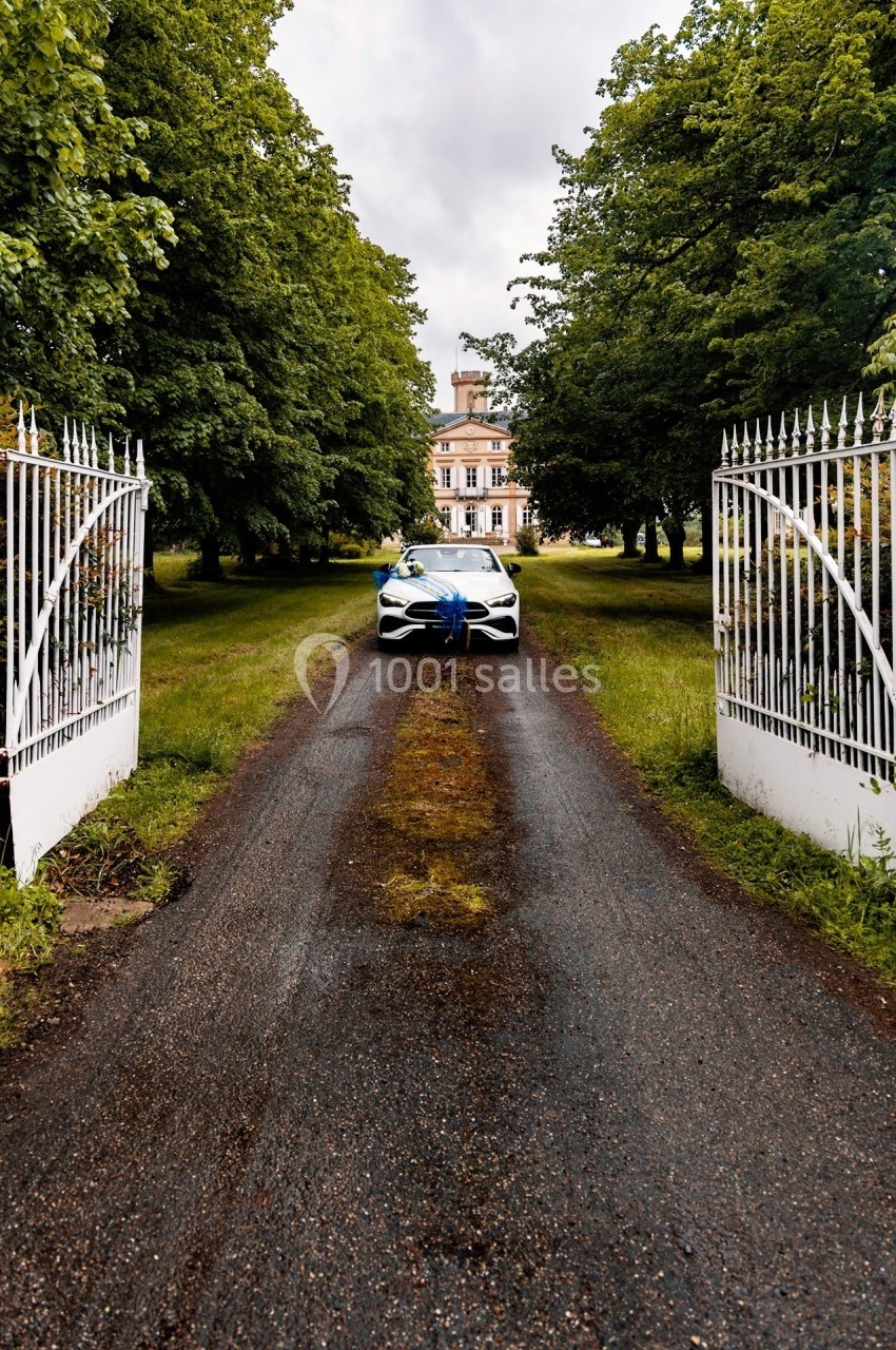Voiture blanche décorée d'un ruban bleu, garée sur une allée bordée d'arbres menant à un manoir.
