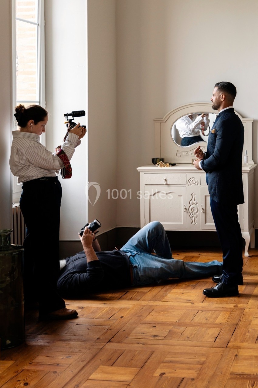 Une femme filme un homme en costume dans une pièce lumineuse, tandis qu'un photographe est allongé au sol.