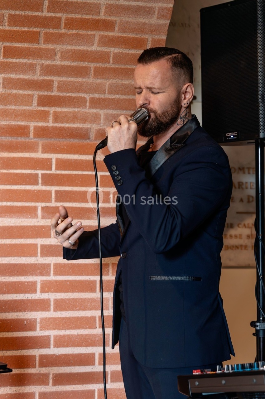 Un homme en costume sombre chante au microphone devant un mur de briques, accompagné d'un équipement audio.